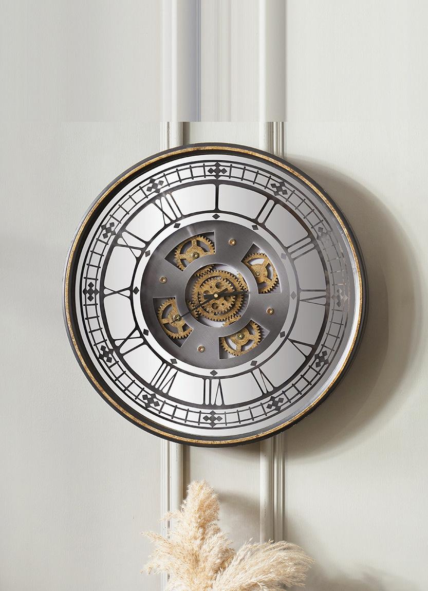 A cog clock with exposed brass gears, hung on a panelled wall above a vase of pampas grass