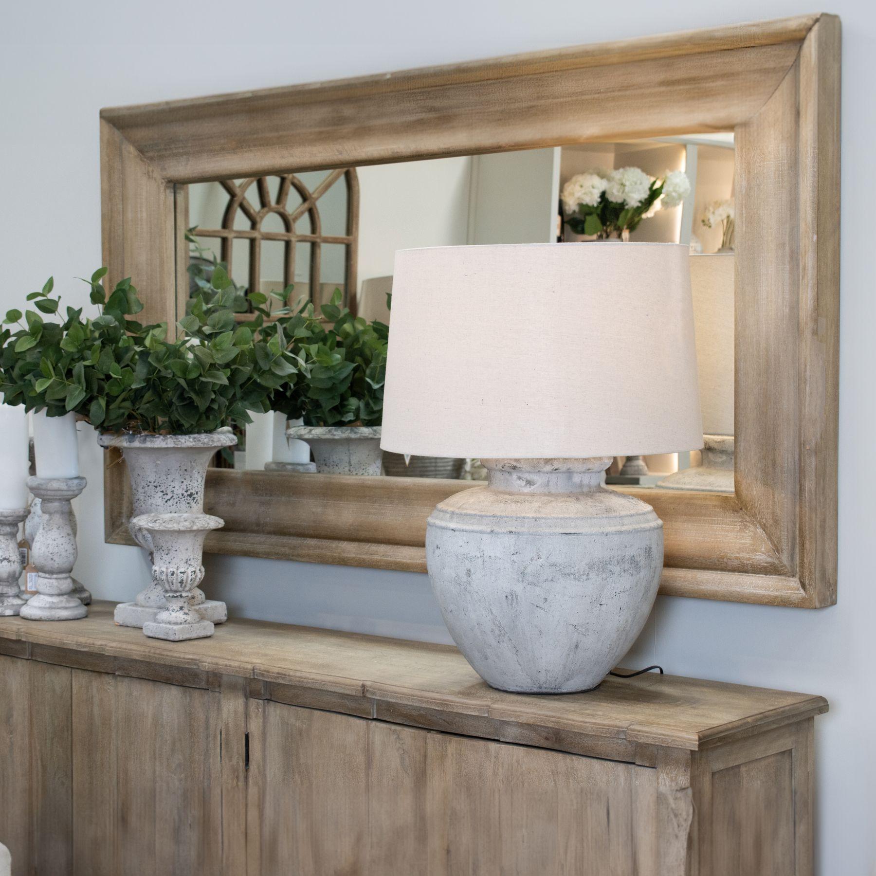 A wood-framed mirror above an oak sideboard styled with a stone urn lamp, eucalyptus and white roses.