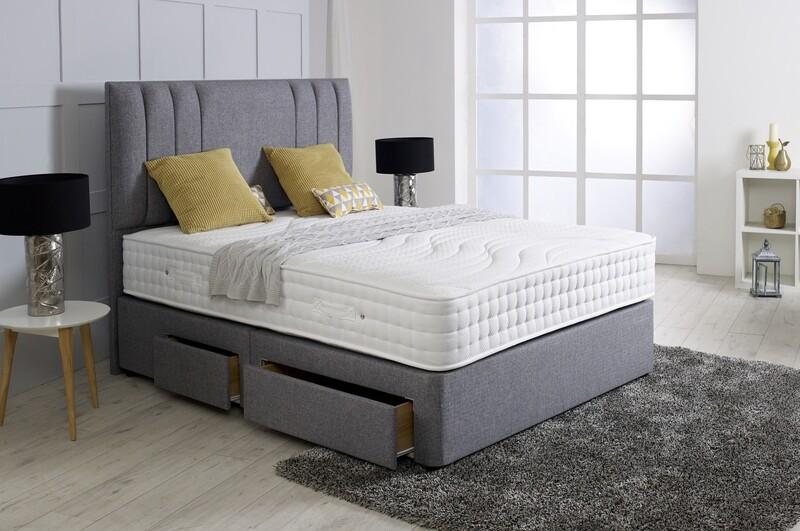 A grey upholstered divan bed with a full headboard and storage drawers, set in a bright bedroom.