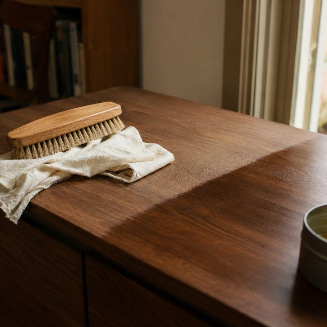 A natural bristle brush and soft cloth on a walnut sideboard beside an open tin of wax.