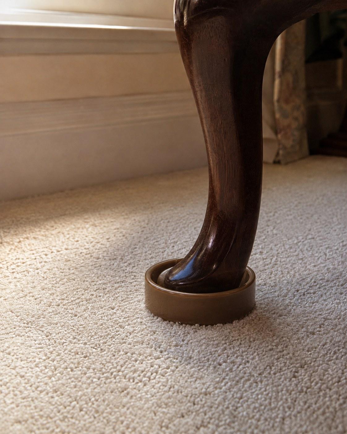The foot of a dark wooden armchair leg resting in a castor cup on a cream wool carpet.