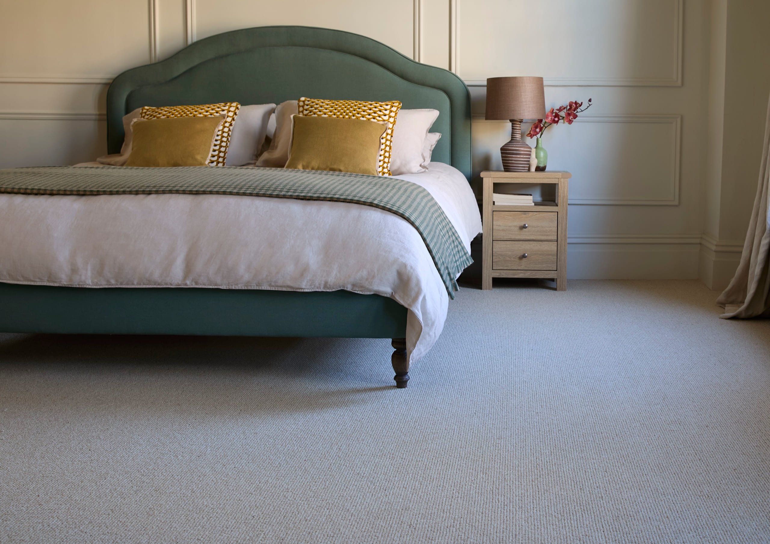 A soft grey Cormar wool carpet running the width of a calm bedroom, with a green upholstered bed and a pale oak bedside table. Flooring by Job's of Oxted