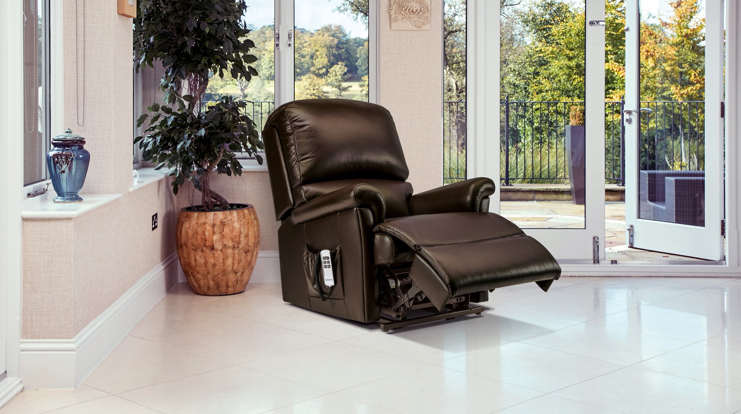A brown leather Sherborne Nevada rise-and-recline chair, reclined with its footrest raised, in a bright garden room