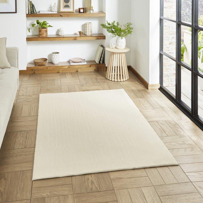 A plain cream rug lying flat on a parquet floor in a bright living room.