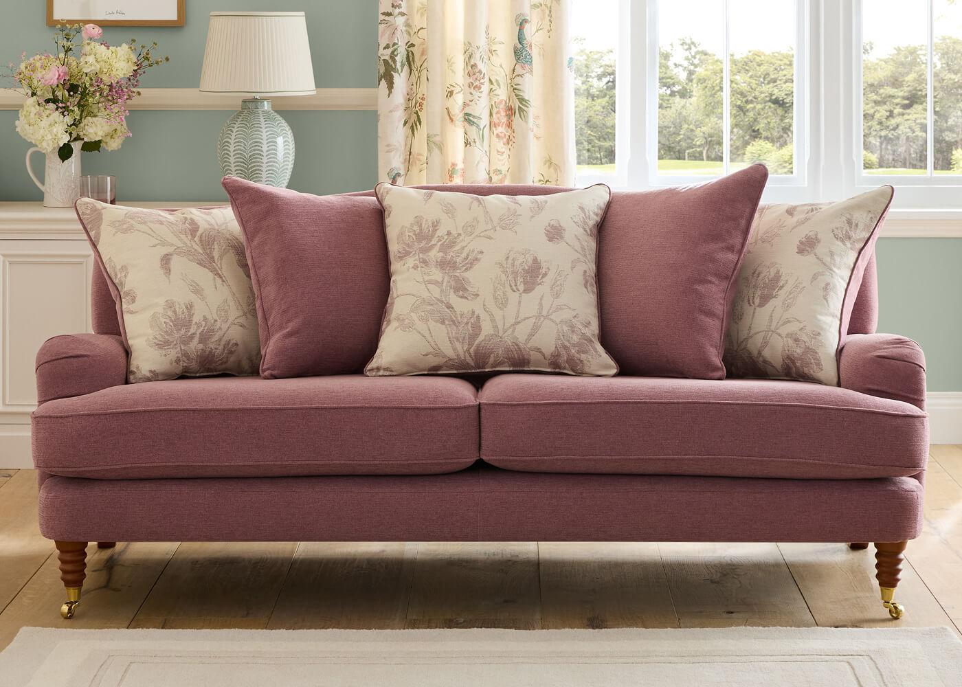 A dusty-pink fabric sofa dressed with floral cushions in a warm, sunlit sitting room